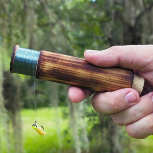 May include: A cylindrical wooden handline fishing reel with a dark stained finish, featuring a cork end cap. The reel is wrapped with thin blue fishing line that extends downward to a small yellow and black fishing lure with a metal hook.