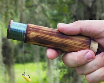Könnte beinhalten: Eine zylindrische Handangelrolle aus Holz mit dunkel gebeizter Oberfläche und einem Kork-Endkappe. Die Rolle ist mit dünner blauer Angelschnur umwickelt, die nach unten zu einem kleinen gelb-schwarzen Angelköder mit Metallhaken führt.