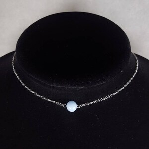 K&ouml;nnte beinhalten: Eine zarte Halskette mit einem kleinen, hellblauen Perlenanh&auml;nger. Die Perle ist auf einer feinen, silberfarbenen Kette zentriert. Die Halskette wird auf einer schwarzen Samtb&uuml;ste pr&auml;sentiert.