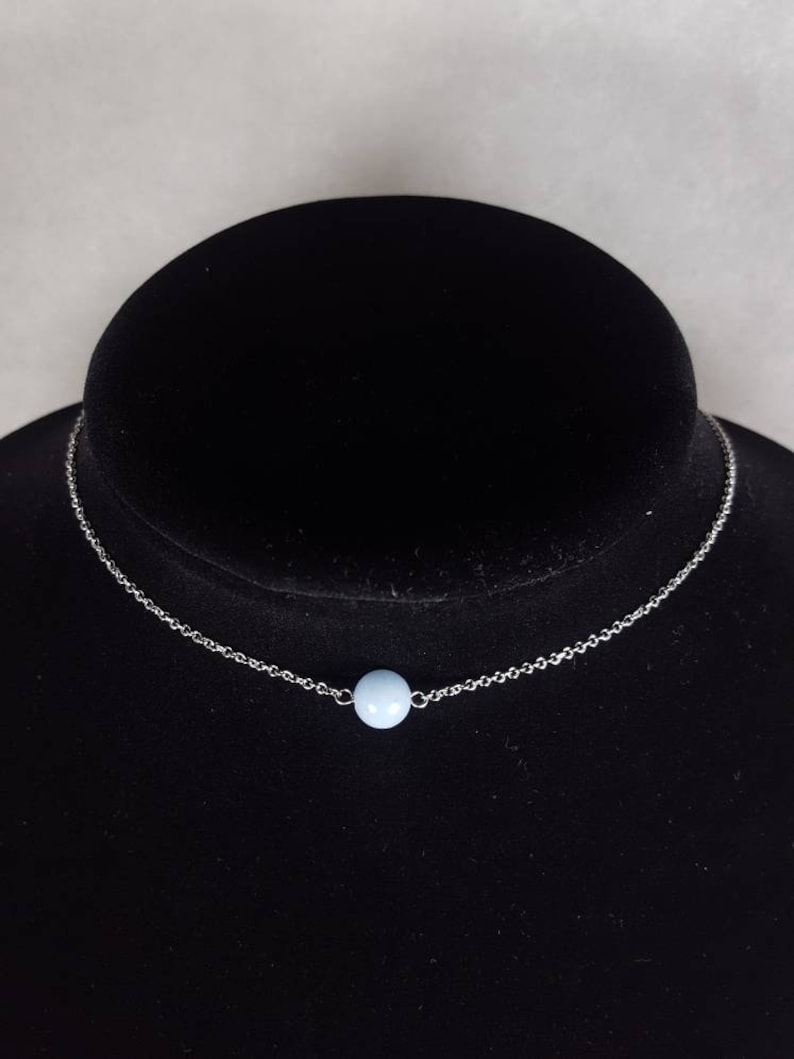 K&ouml;nnte beinhalten: Eine zarte Halskette mit einem kleinen, hellblauen Perlenanh&auml;nger. Die Perle ist auf einer feinen, silberfarbenen Kette zentriert. Die Halskette wird auf einer schwarzen Samtb&uuml;ste pr&auml;sentiert.