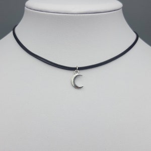 May include: A simple silver crescent moon charm hangs from a black choker necklace.