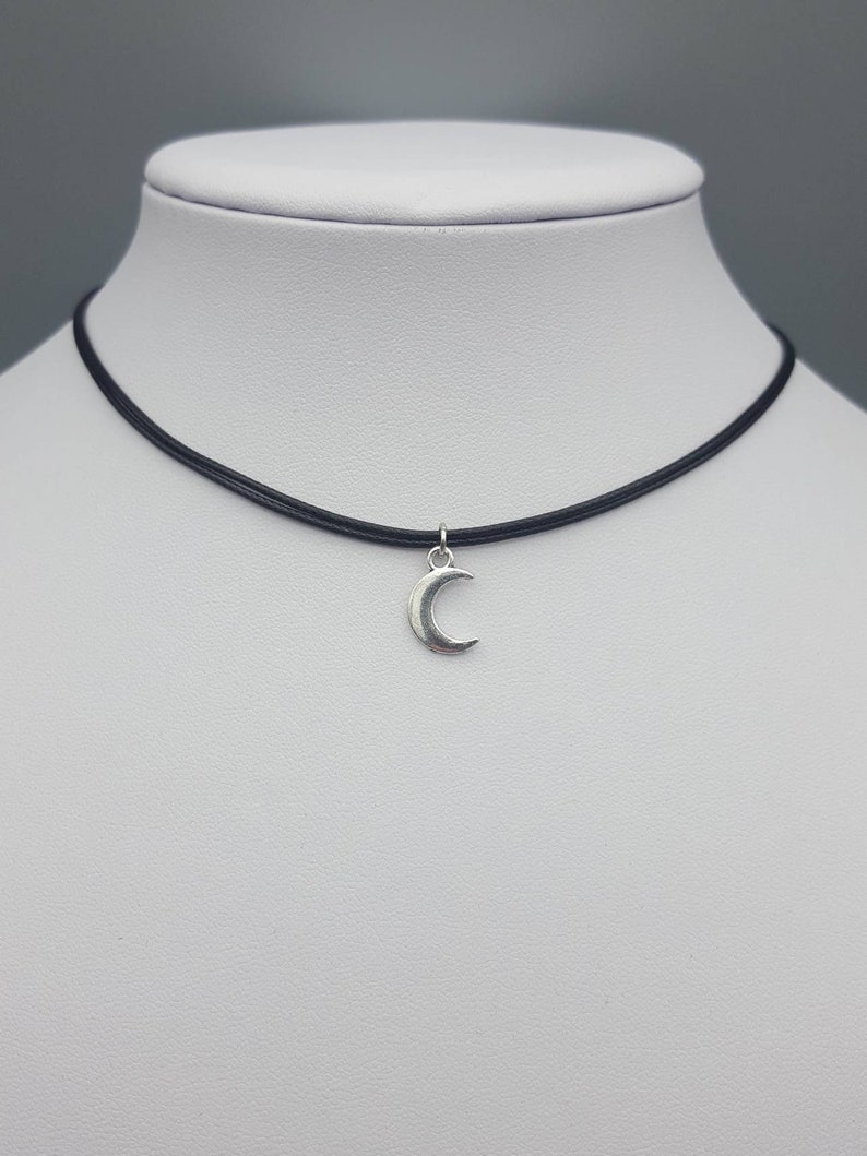 May include: A simple silver crescent moon charm hangs from a black choker necklace.
