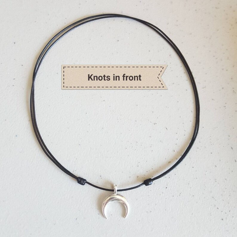 May include: A black cord necklace with a silver crescent moon pendant. The necklace has knots at the front.
