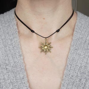 May include: A bronze-colored sun pendant necklace with a black cord. The sun pendant has a detailed, radiating design with a central circular element. The necklace is worn around the neck.