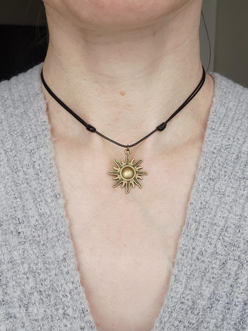 May include: A bronze-colored sun pendant necklace with a black cord. The sun pendant has a detailed, radiating design with a central circular element. The necklace is worn around the neck.