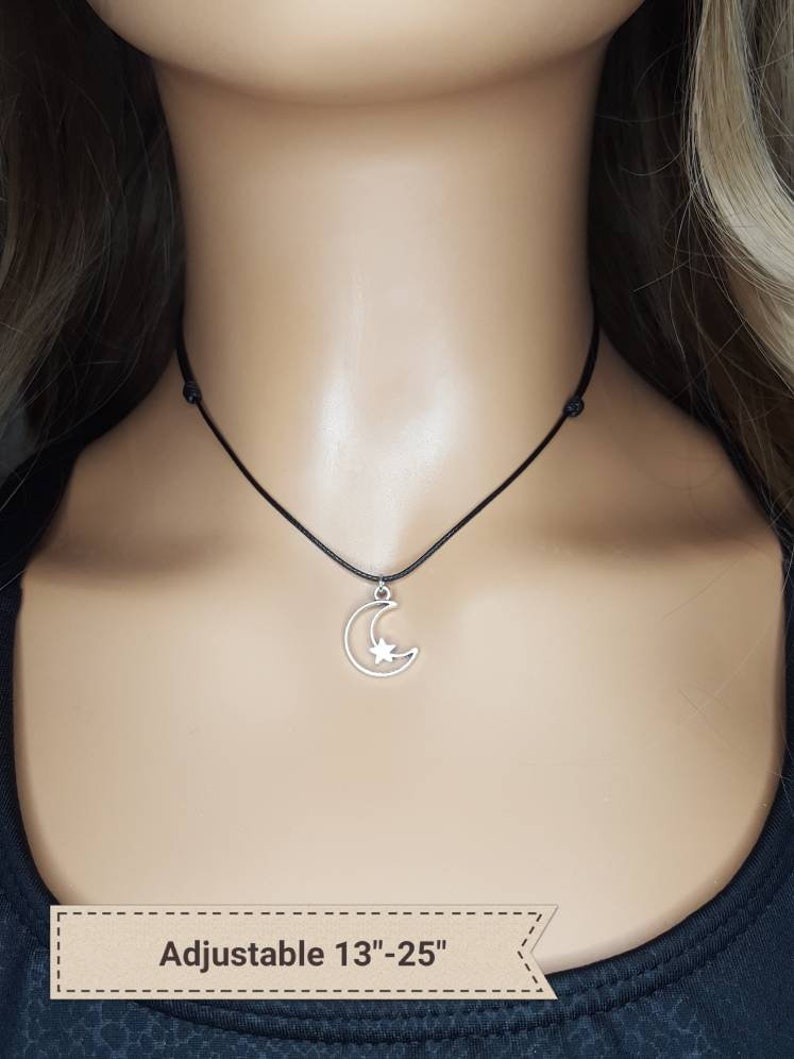 May include: A silver crescent moon charm with a star inside hangs from a black adjustable cord necklace. The necklace is adjustable from 13 to 25 inches.