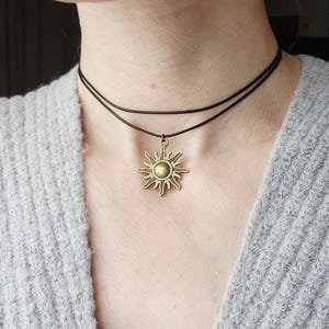 May include: A layered choker necklace featuring a bronze sun pendant. The necklace has two thin, dark brown cords. The sun pendant has a detailed design with rays extending outward from a central circular element. The necklace is worn against a gray sweater.