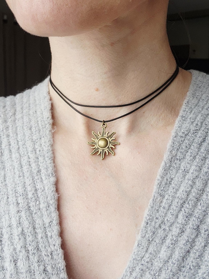 May include: A layered choker necklace featuring a bronze sun pendant. The necklace has two thin, dark brown cords. The sun pendant has a detailed design with rays extending outward from a central circular element. The necklace is worn against a gray sweater.