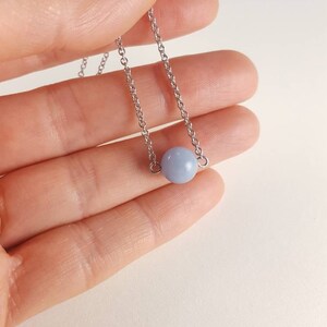 K&ouml;nnte beinhalten: Eine zarte Halskette mit einem hellblauen Perlenanh&auml;nger. Die Perle h&auml;ngt an einer feinen, silberfarbenen Kette. Die Halskette wird in einer Hand vor einem weichen, neutralen Hintergrund gehalten.