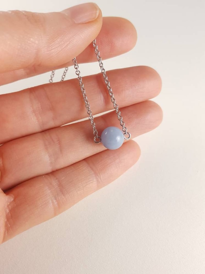 K&ouml;nnte beinhalten: Eine zarte Halskette mit einem hellblauen Perlenanh&auml;nger. Die Perle h&auml;ngt an einer feinen, silberfarbenen Kette. Die Halskette wird in einer Hand vor einem weichen, neutralen Hintergrund gehalten.