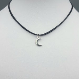 May include: A silver crescent moon charm hangs from a black leather choker necklace.