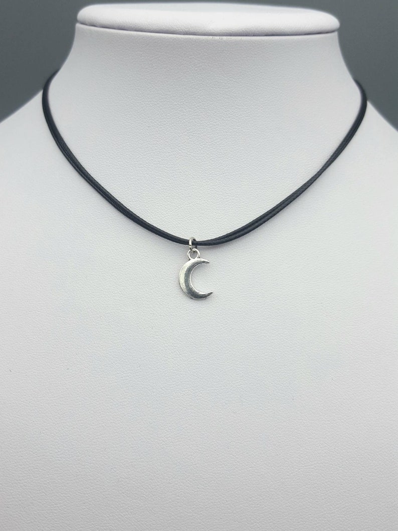 May include: A silver crescent moon charm hangs from a black leather choker necklace.