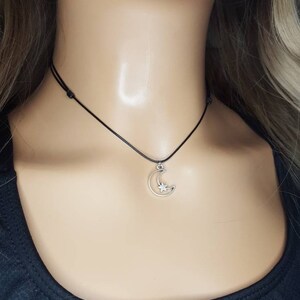 May include: A silver crescent moon charm necklace with a small star inside the moon. The necklace is on a black cord.