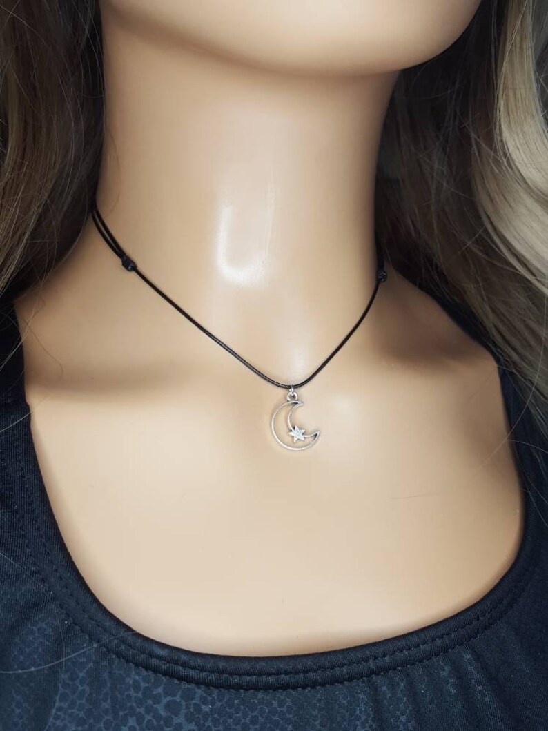 May include: A silver crescent moon charm necklace with a small star inside the moon. The necklace is on a black cord.
