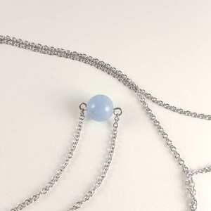 K&ouml;nnte beinhalten: Eine zarte Halskette mit einem hellblauen, kugelf&ouml;rmigen Perlenanh&auml;nger. Der Anh&auml;nger ist an einer feinen, silberfarbenen Kette befestigt. Die Halskette hat ein mehrschichtiges Design mit mehreren Kettengliedern.