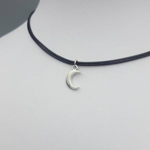 May include: A silver crescent moon charm hangs from a black choker necklace. The necklace is made of thin, black cord and is adjustable.
