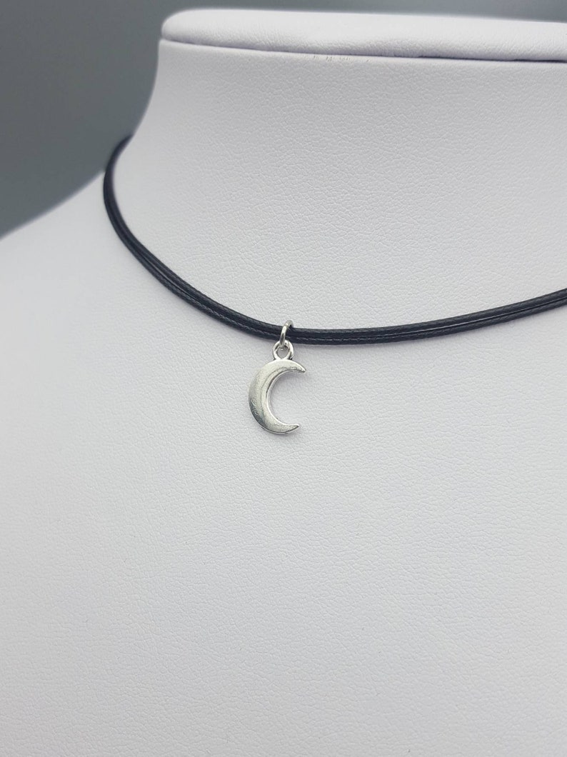 May include: A silver crescent moon charm hangs from a black choker necklace. The necklace is made of thin, black cord and is adjustable.