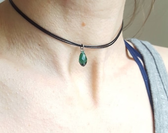 May include: A black choker necklace with a small, teardrop-shaped, emerald green crystal pendant. The necklace is worn around the neck, and the pendant hangs in the center. The crystal is faceted, catching the light.