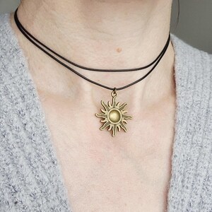 May include: A layered black choker necklace with a bronze sun pendant. The sun pendant has a detailed design with rays extending outward from a central circle. The necklace is worn against a gray sweater.