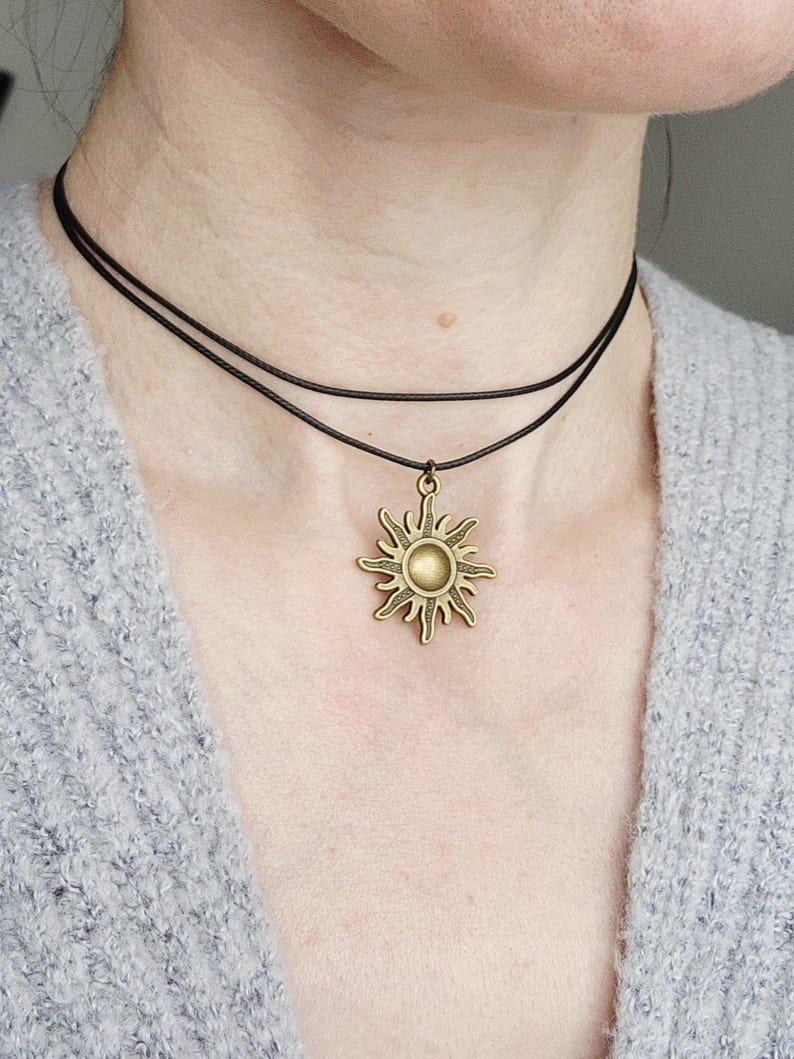May include: A layered black choker necklace with a bronze sun pendant. The sun pendant has a detailed design with rays extending outward from a central circle. The necklace is worn against a gray sweater.