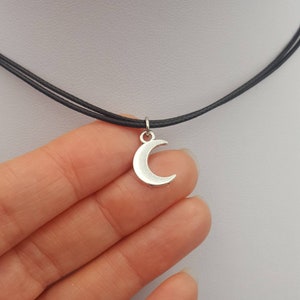 May include: A silver crescent moon charm hangs from a black choker necklace.