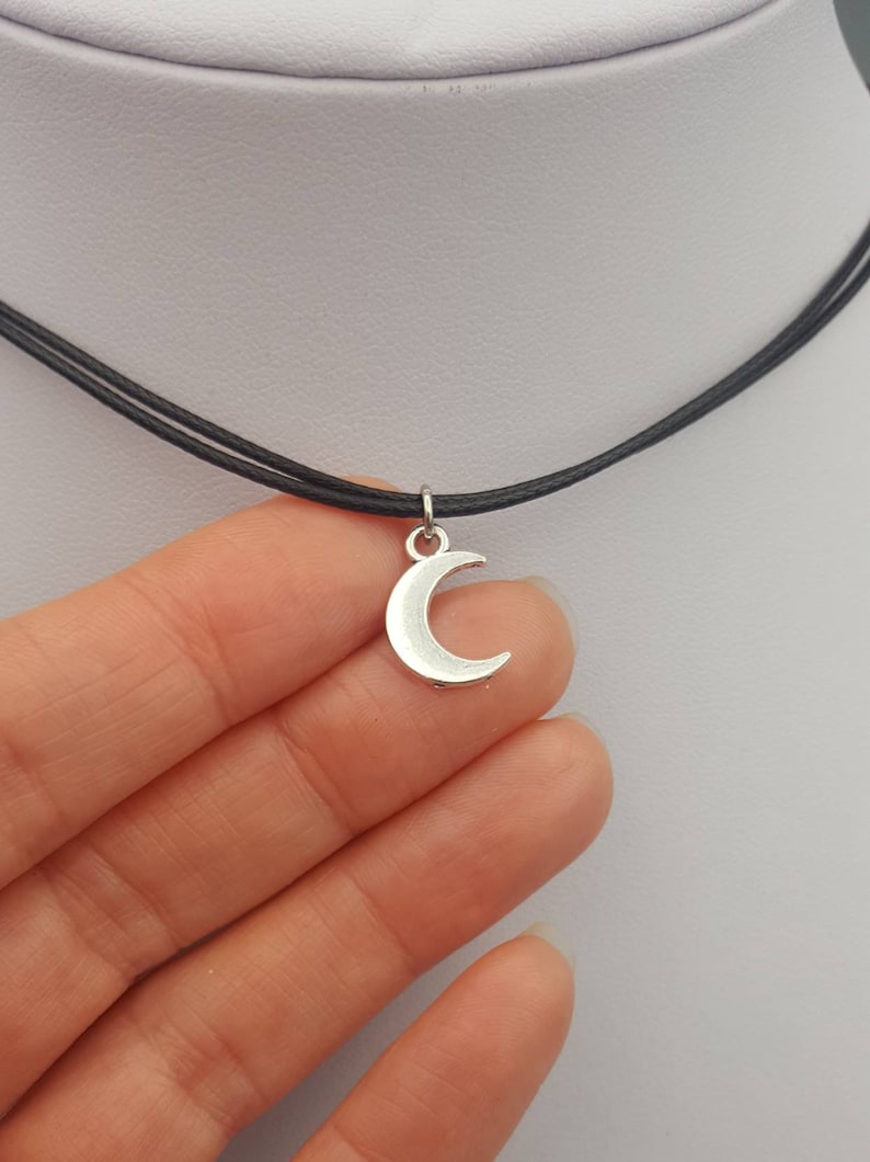 May include: A silver crescent moon charm hangs from a black choker necklace.