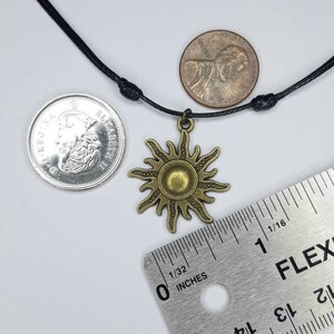 May include: A bronze-colored sun pendant necklace with a black cord. The sun pendant has a detailed design with rays extending outward. A ruler shows inches. A penny and a coin are next to the necklace.