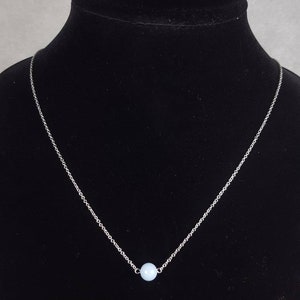 K&ouml;nnte beinhalten: Eine silberne Halskette mit einem hellblauen Perlenanh&auml;nger. Die Halskette wird auf einer schwarzen Schaufensterpuppe pr&auml;sentiert. Die Kette ist fein und die Perle ist rund. Das Design ist minimalistisch und unterstreicht die sanfte Farbe der Perle.