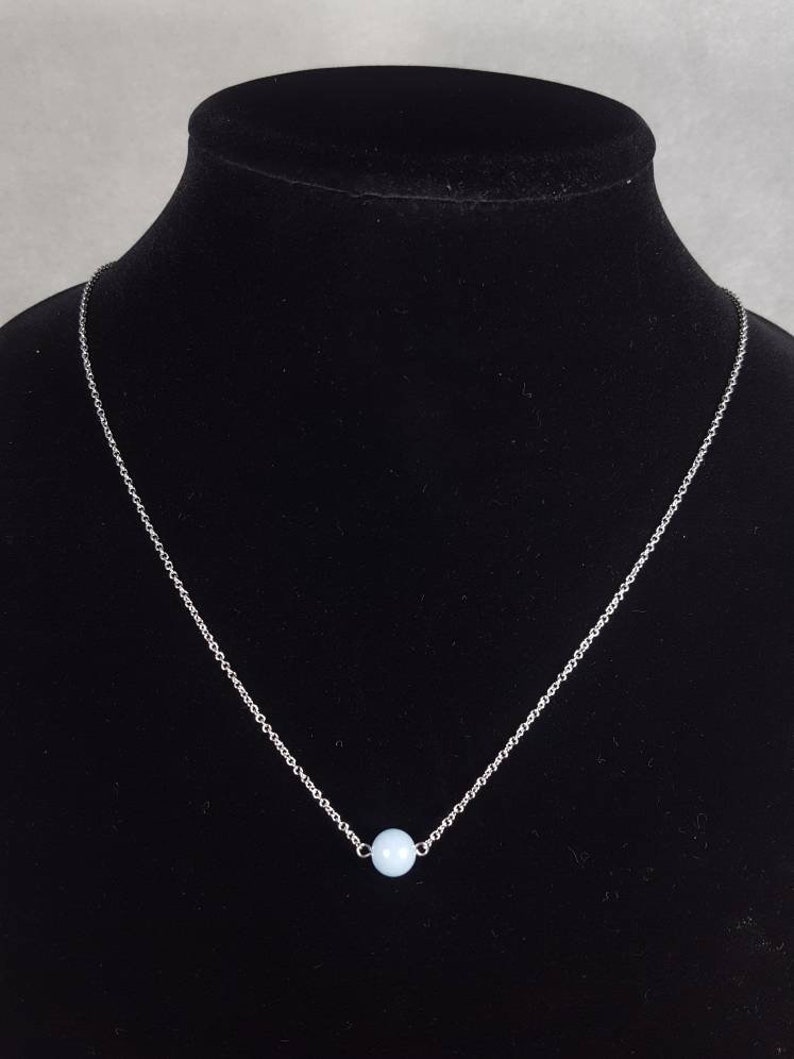 K&ouml;nnte beinhalten: Eine silberne Halskette mit einem hellblauen Perlenanh&auml;nger. Die Halskette wird auf einer schwarzen Schaufensterpuppe pr&auml;sentiert. Die Kette ist fein und die Perle ist rund. Das Design ist minimalistisch und unterstreicht die sanfte Farbe der Perle.