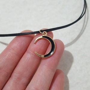 May include: A black choker necklace with a gold crescent moon pendant. The moon is black with a gold outline.
