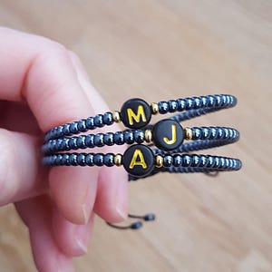 May include: Three dark grey beaded bracelets, each with a letter: "M", "J", and "A" on black and gold beads. The bracelets have gold accents and a black adjustable closure.