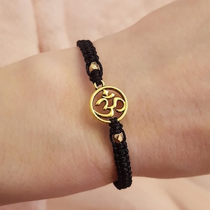 May include: Black woven bracelet featuring a gold-colored Om symbol charm. The charm is circular with an intricate design. The bracelet has gold-colored beads and is worn on a wrist.