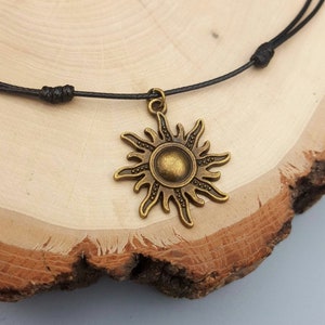 May include: A bronze-colored sun pendant necklace with a black cord. The sun pendant has a detailed design with rays and a central circular element. The necklace is displayed on a wooden surface.