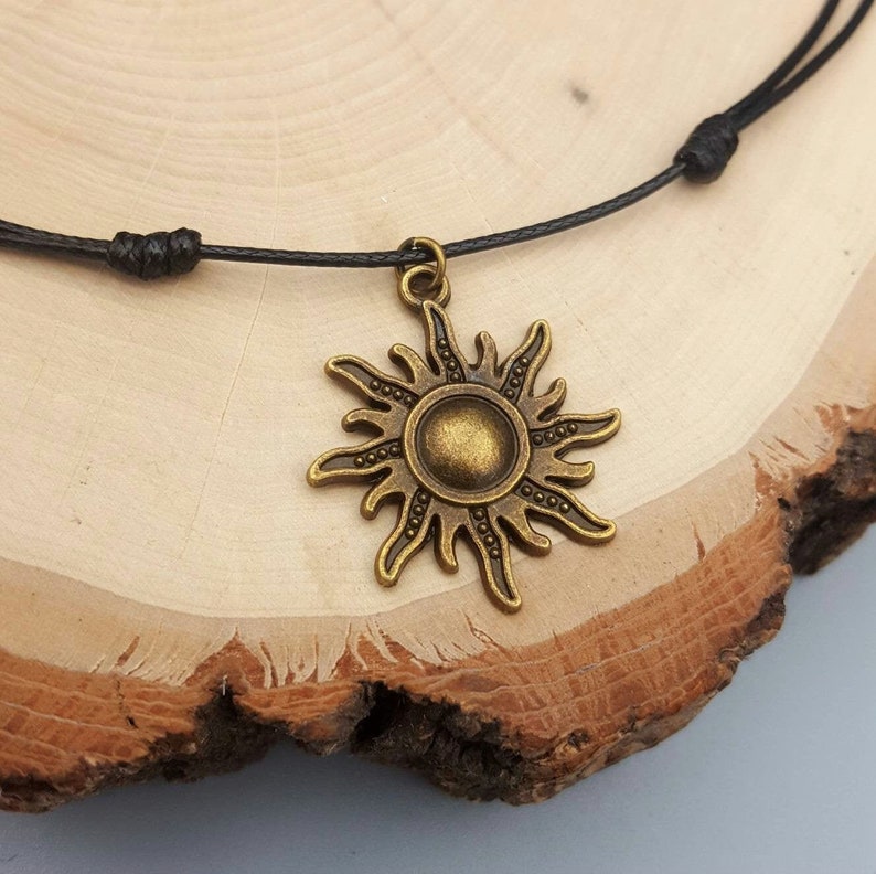 May include: A bronze-colored sun pendant necklace with a black cord. The sun pendant has a detailed design with rays and a central circular element. The necklace is displayed on a wooden surface.