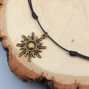 May include: A bronze-colored sun pendant necklace with a black cord. The sun pendant has detailed rays and a circular center. The necklace is displayed on a wooden surface.