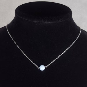 K&ouml;nnte beinhalten: Eine zarte Silberkette mit einem kleinen, hellblauen Perlenanh&auml;nger. Die Halskette wird auf einer schwarzen Schaufensterpuppe pr&auml;sentiert, die das minimalistische Design und den subtilen Farbkontrast hervorhebt.