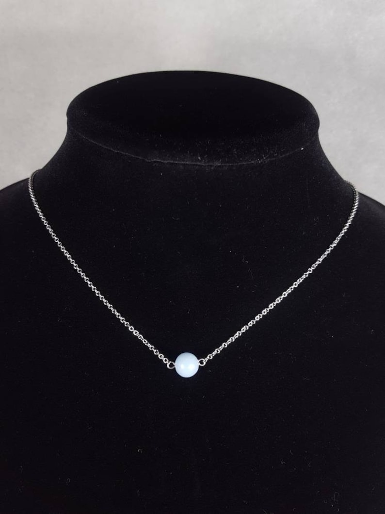 K&ouml;nnte beinhalten: Eine zarte Silberkette mit einem kleinen, hellblauen Perlenanh&auml;nger. Die Halskette wird auf einer schwarzen Schaufensterpuppe pr&auml;sentiert, die das minimalistische Design und den subtilen Farbkontrast hervorhebt.