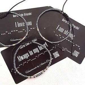 Könnte beinhalten: Drei Armbänder mit Perlen, schwarzen Kordeln und silbernen Perlen. Jedes Armband wird auf einer schwarzen Karte mit weißem Text präsentiert. Die Karten zeigen "I love you", "I am strong" und "Always in my heart" in Morsecode.