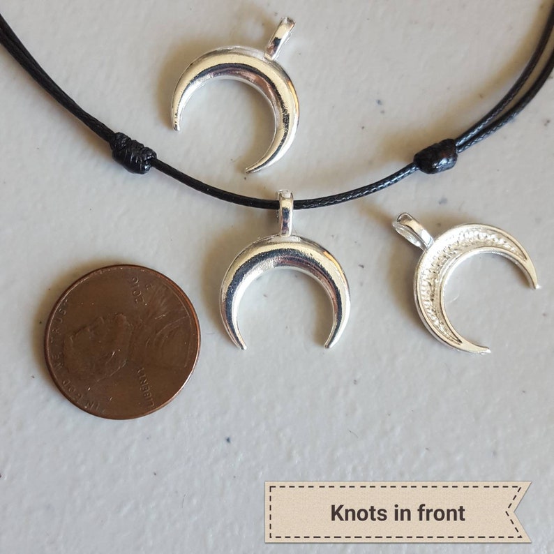 May include: Three silver crescent moon charms on a black cord necklace. The necklace has knots at the front. The charms are about the size of a UK penny.