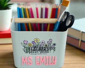 Puede incluir: Un organizador de escritorio azul claro con asa de madera, lleno de lápices de colores, pinceles y tijeras. La parte delantera presenta el texto "TEACHER'S plant seeds that grow forever" y el nombre "MS EMILY" en rosa.