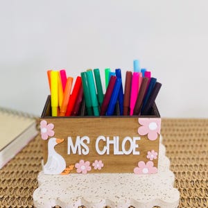 May include: A rectangular wooden desk organiser filled with various colourful markers. The front features a white goose illustration, the text "MS CHLOE" in white lettering, and several light pink flower cutouts attached to the wood surface.
