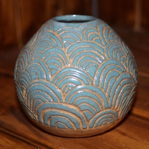 May include: A small, round ceramic vase with a light blue glaze and a repeating wave pattern in a lighter tan color. The vase has a wide opening and sits on a tan base. The design is carved into the surface.