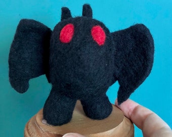 May include: A black felt plush toy shaped like a bat with large red eyes. The toy is standing on a wooden base.