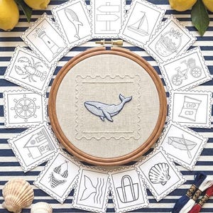 Può includere: Un telaio da ricamo in legno contenente un pannello di tessuto beige con un ricamo di balena blu, circondato da una disposizione circolare di carte di carta bianca con disegni a linee nere di icone nautiche e un sacchetto di patatine.