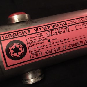 May include: A pink label with black text and a black Imperial symbol. The label has the text "12000WF47", "VIDE", "M7", "74710", "VOLTE", "70X7", "K۷۱۲M 0047", "07KYW 40417207", "CO", "MOOVIC RX", "ZAVOD", "4152728", "701M", "LAST", "V", "ИМЯ ПО ЛИNDOLT AS", "12000WF47", "7M037".