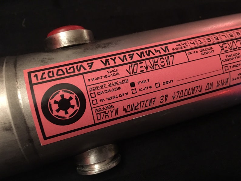 May include: A pink label with black text and a black Imperial symbol. The label has the text "12000WF47", "VIDE", "M7", "74710", "VOLTE", "70X7", "K۷۱۲M 0047", "07KYW 40417207", "CO", "MOOVIC RX", "ZAVOD", "4152728", "701M", "LAST", "V", "ИМЯ ПО ЛИNDOLT AS", "12000WF47", "7M037".