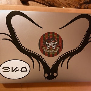 Puede incluir: Un port&aacute;til plateado con una pegatina negra de una pareja de cuernos de ciervo. La pegatina tiene forma de coraz&oacute;n. Hay una pegatina redonda con un dise&ntilde;o de calavera y huesos cruzados en el port&aacute;til. La pegatina dice "The Boozehounds Live Experience". Hay una pegatina ovalada blanca con el texto "EKO" en el port&aacute;til.