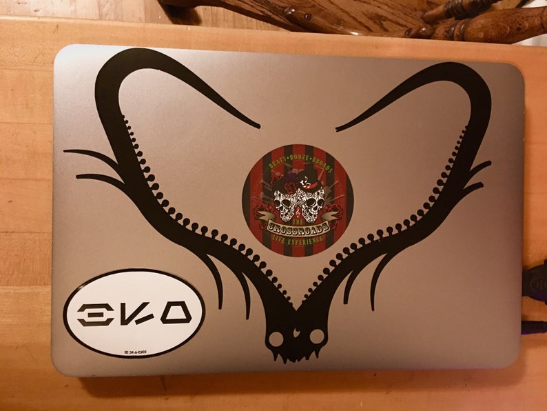 Puede incluir: Un port&aacute;til plateado con una pegatina negra de una pareja de cuernos de ciervo. La pegatina tiene forma de coraz&oacute;n. Hay una pegatina redonda con un dise&ntilde;o de calavera y huesos cruzados en el port&aacute;til. La pegatina dice "The Boozehounds Live Experience". Hay una pegatina ovalada blanca con el texto "EKO" en el port&aacute;til.