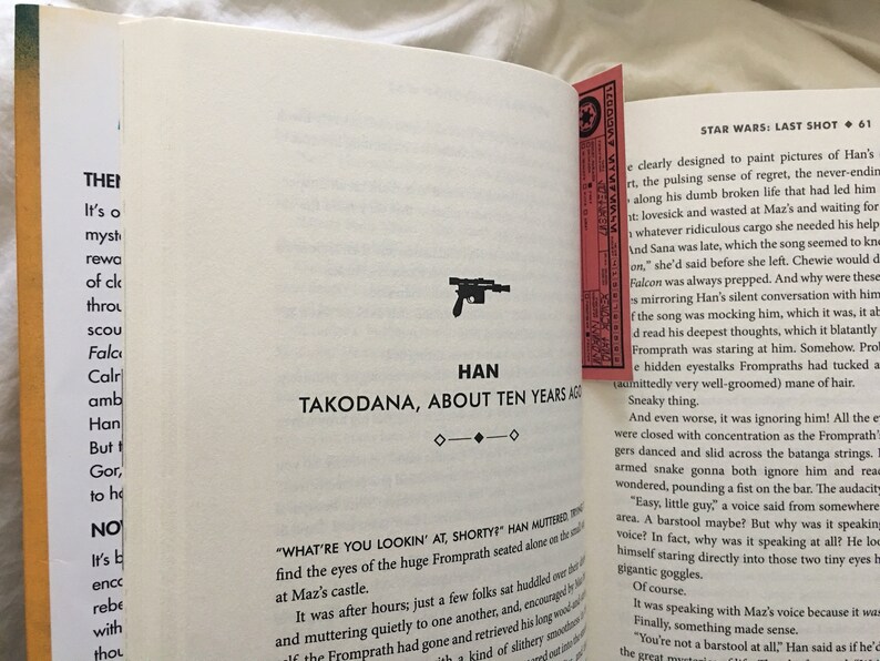 May include: A close-up of a book page with text about Star Wars: Last Shot. The text describes Han Solo's thoughts and feelings as he is on the planet Takodana. The page also features a small illustration of a blaster.