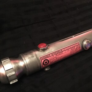 May include: A silver lightsaber hilt with a red button and a black and white sticker with the text "120007  KYU7F1M1M  V02-53627".